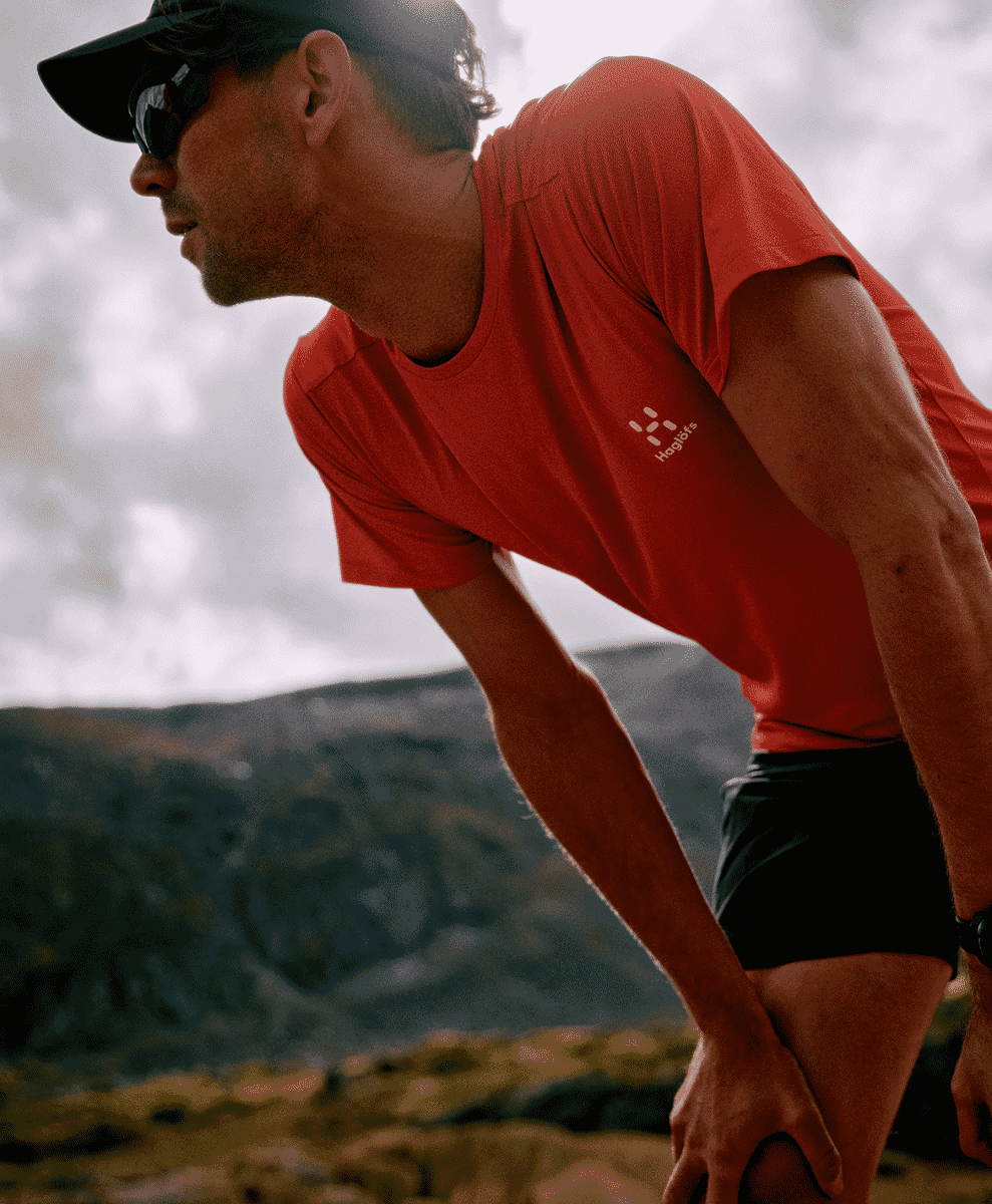 A male runner in a red performance t-shirt and black shorts standing on a rugged mountain trail.
