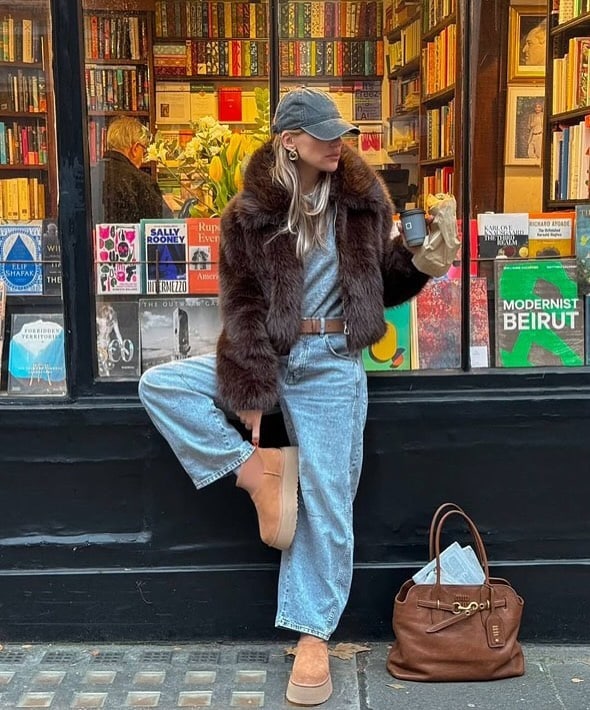 Woman in a brown faux fur coat and blue jeans holding coffee outside a bookstore.