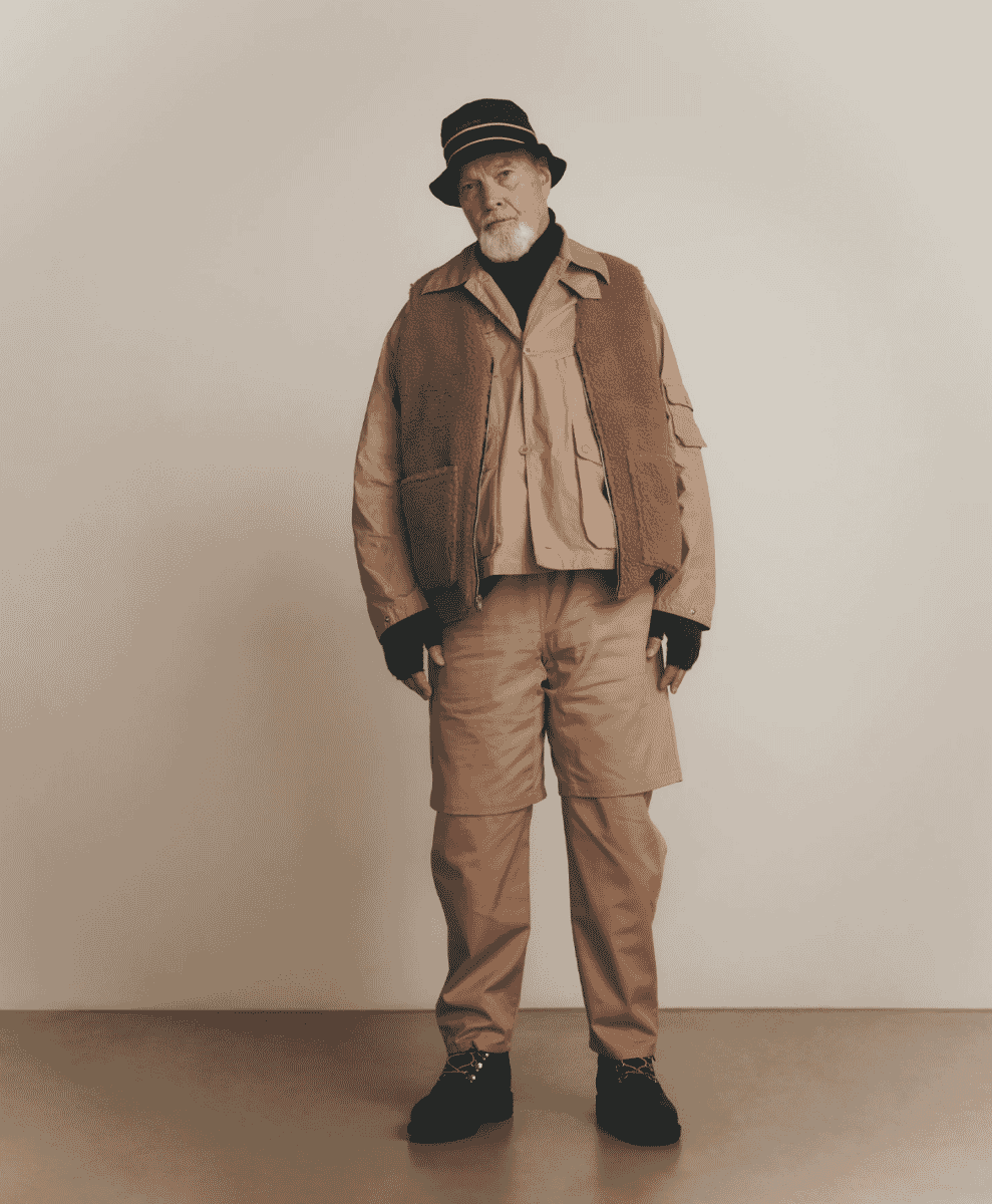 Older man with a white beard wearing a layered shearling vest and beige utility jacket.