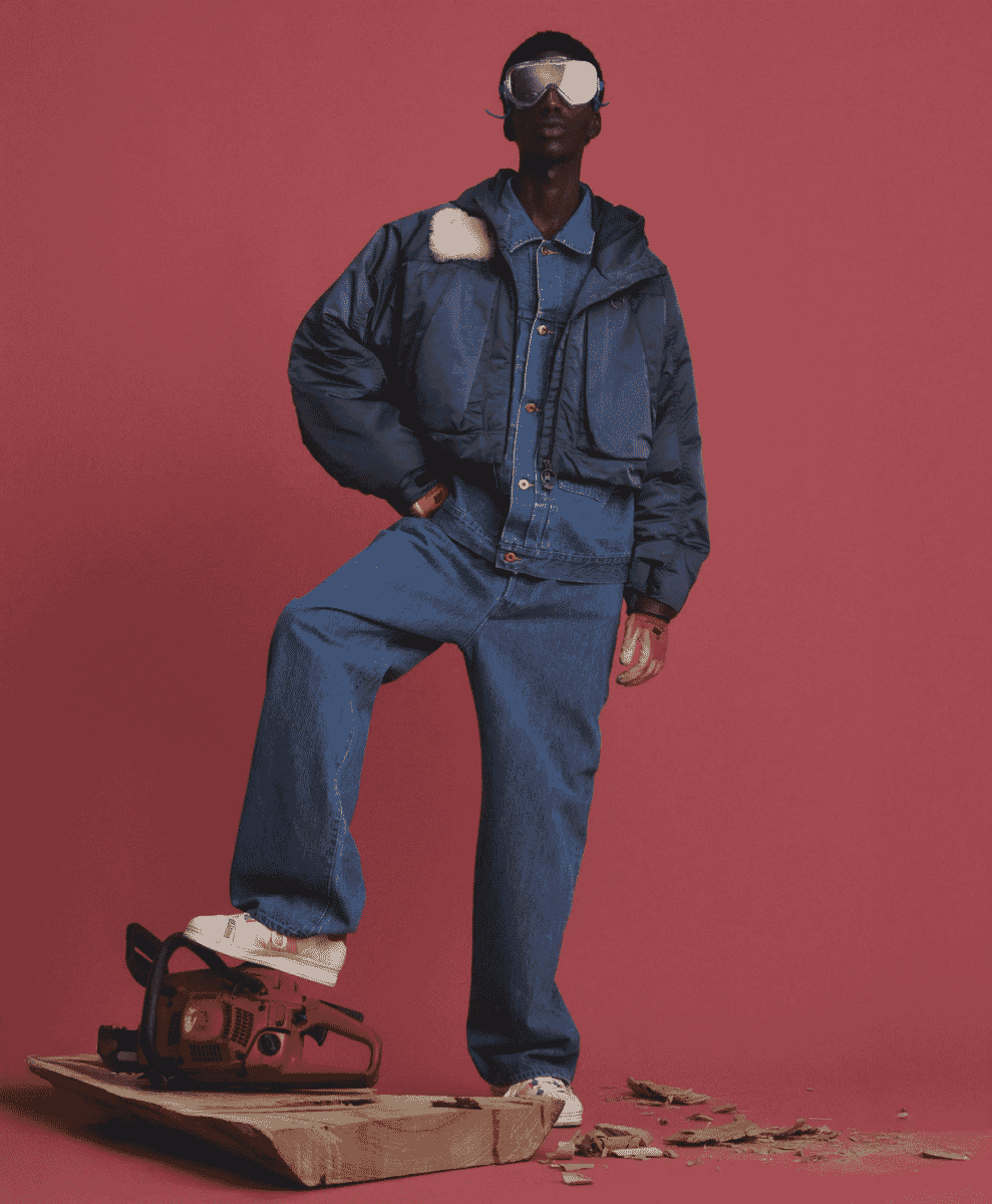 Model in a blue denim puffer jacket and ski goggles standing on a cut log.