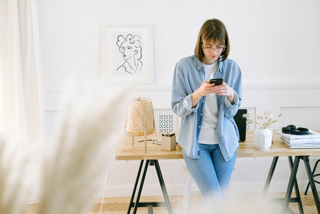 Woman in denim-on-denim outfit browsing her phone at a minimalist wooden desk with soft lighting.