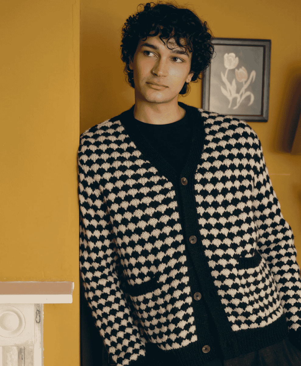 A man in a black and ecru houndstooth cardigan over a black turtleneck against a mustard-yellow wall.