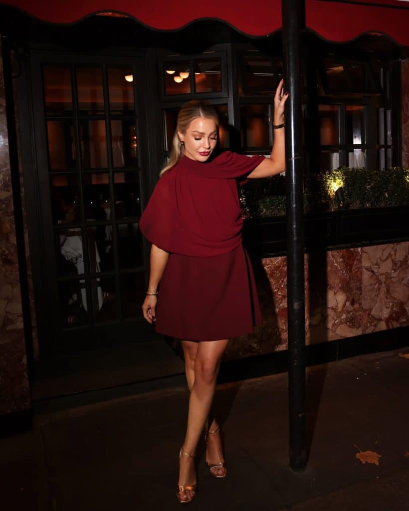 Woman wearing a burgundy draped mini dress and gold heels outside a restaurant at night.