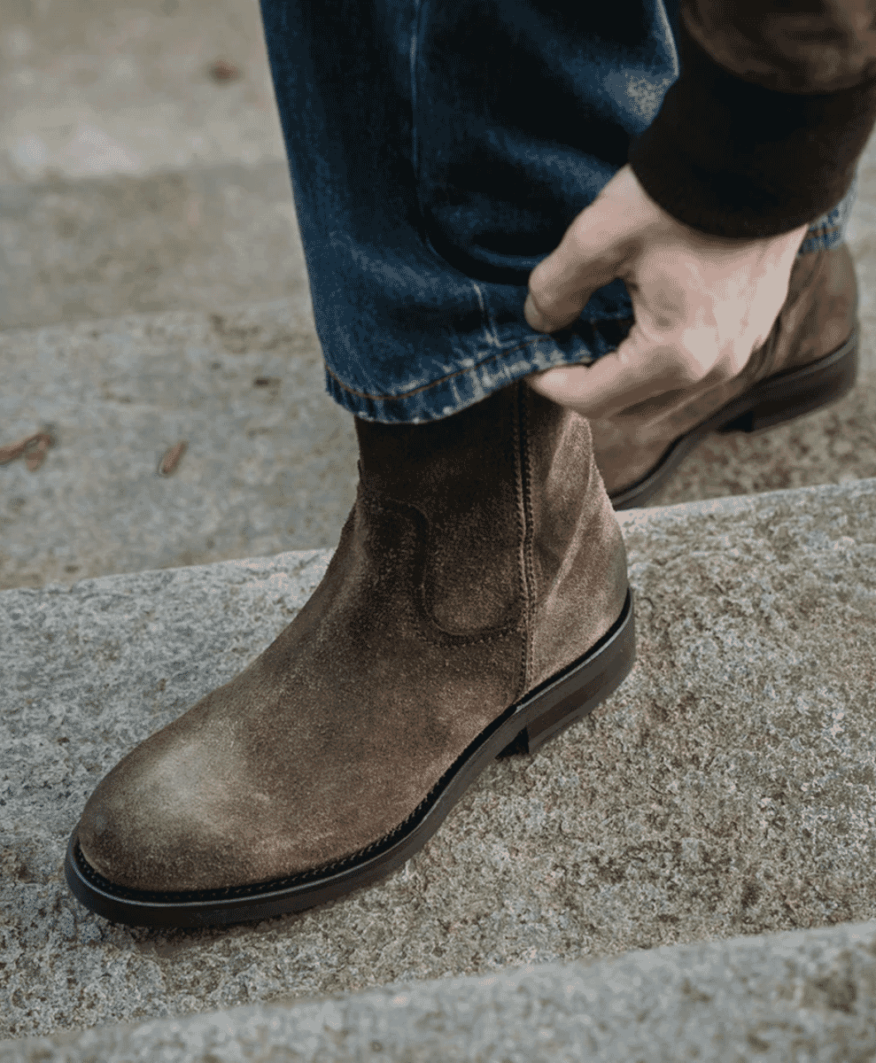 Close-up of a man adjusting blue jeans over brown suede Chelsea boots.