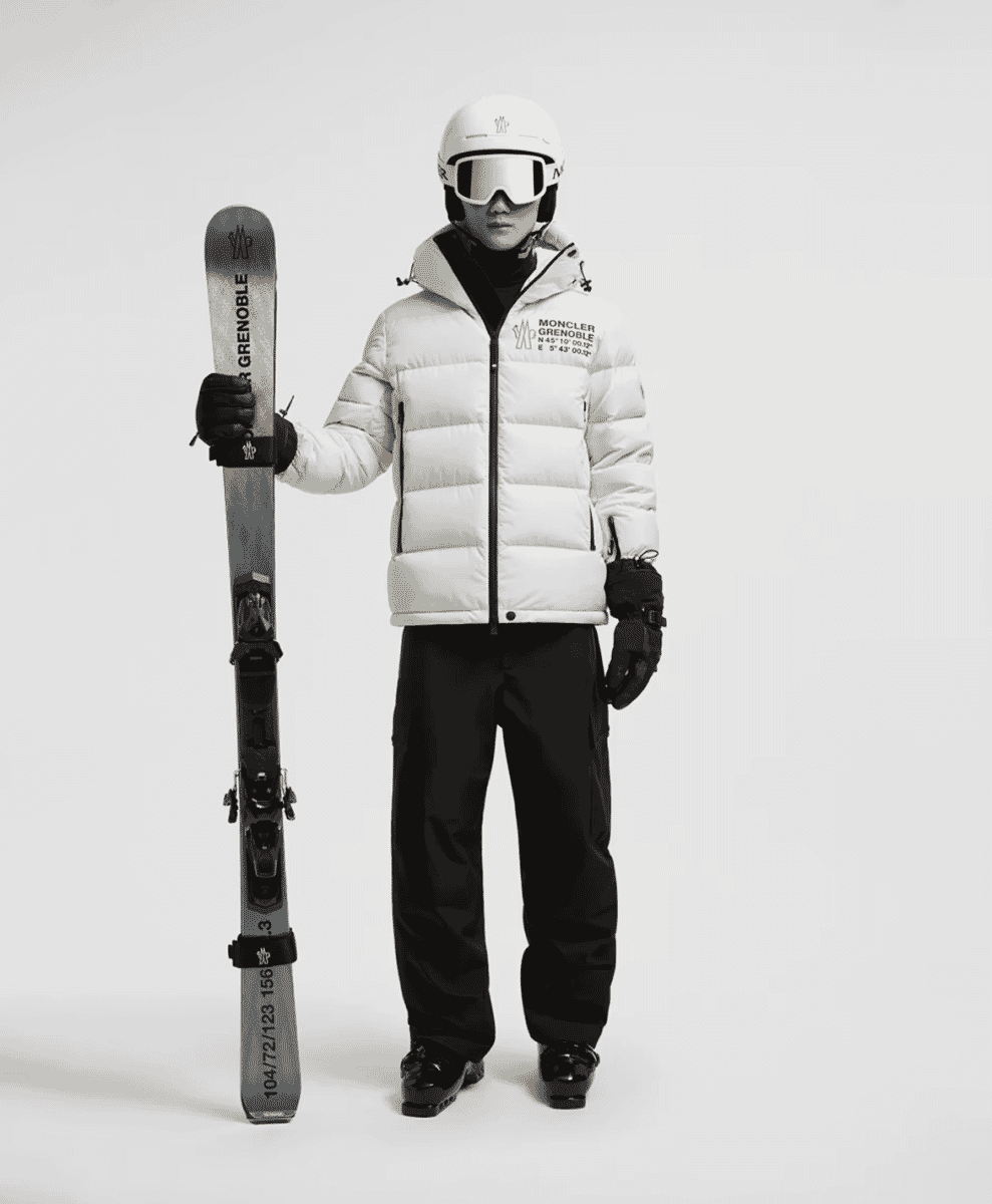 A model in a white Moncler Grenoble puffer jacket and black pants holding a ski against a white background.