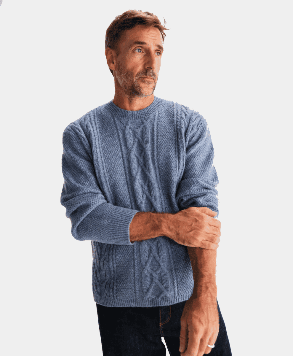 A man in a blue cable-knit sweater and dark jeans standing against a plain studio background.
