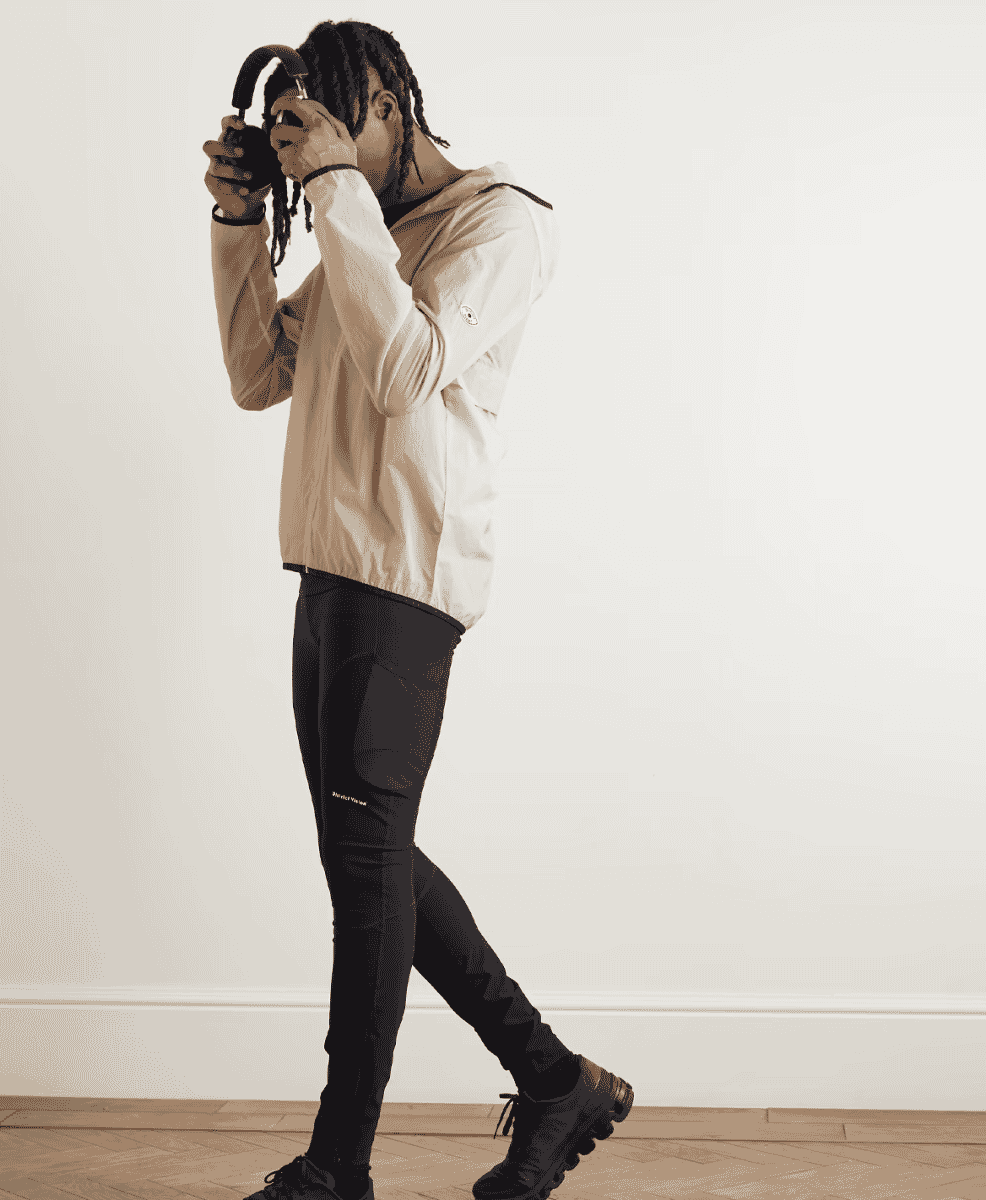 A man wearing a light beige hooded jacket and black compression pants against a white studio background.