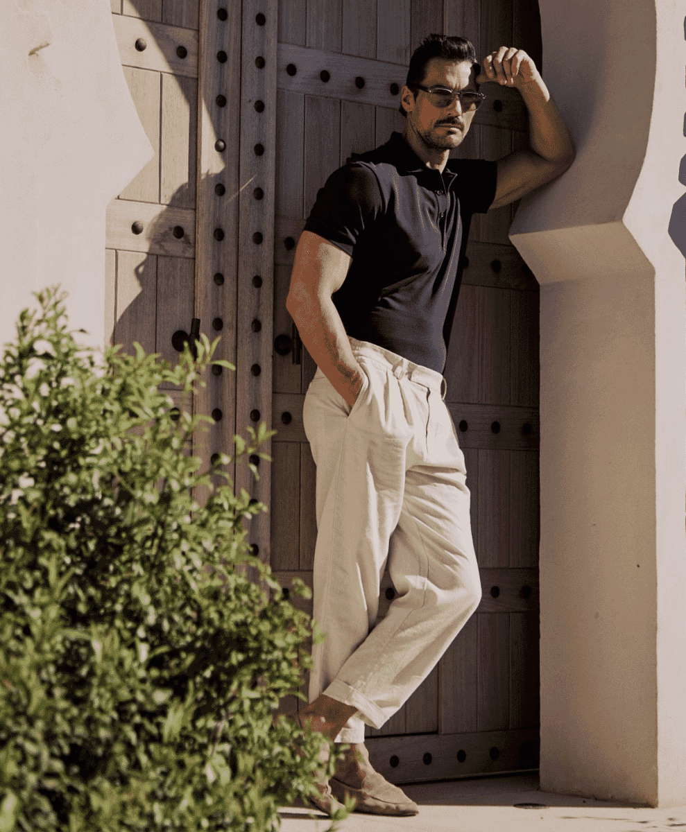 Man wearing cream-colored wide-leg trousers and a black polo leaning against a wooden door.