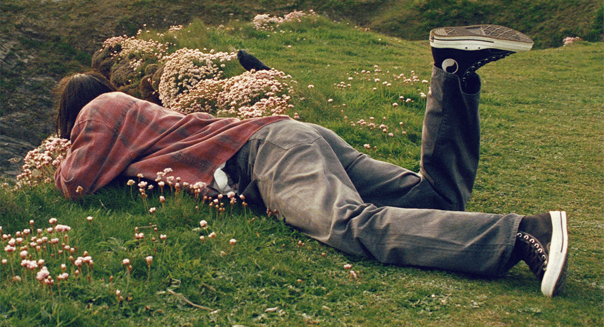 A person lying in a meadow wearing a plaid shirt and black Converse sneakers.