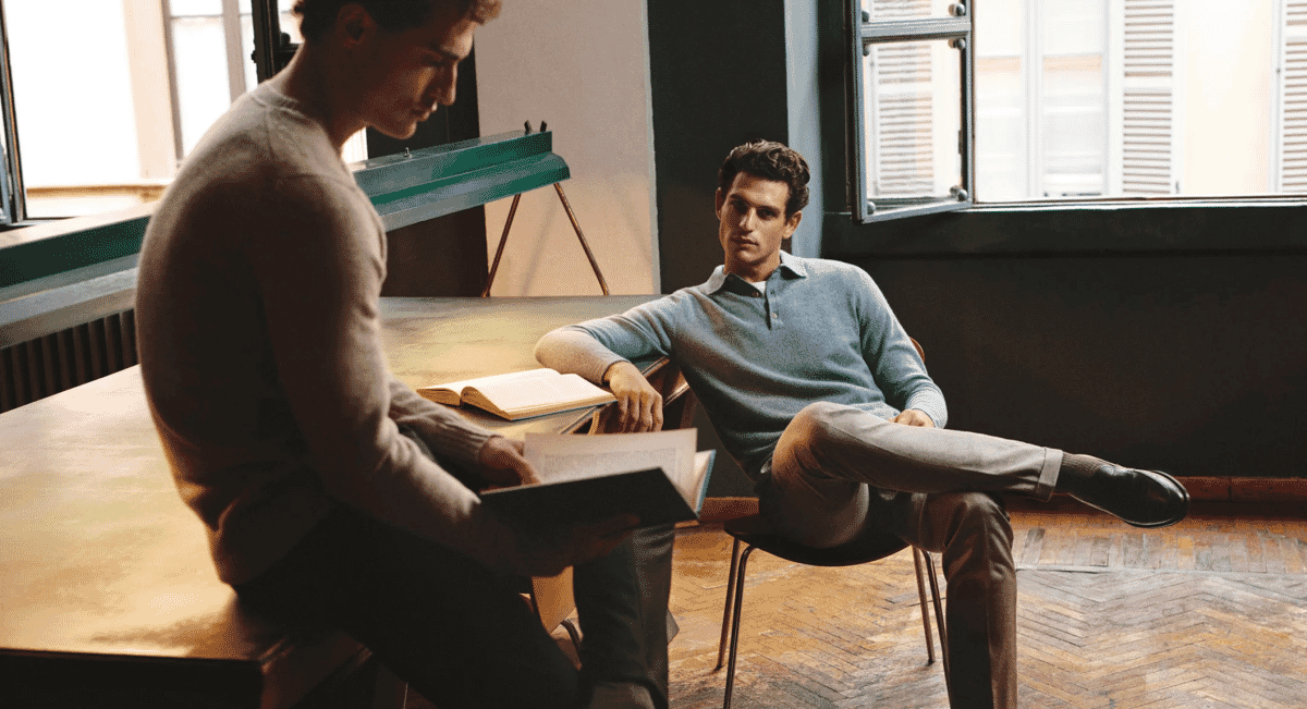 Two men in an intellectual indoor setting wearing a beige sweater and a light blue polo knit with tailored trousers.