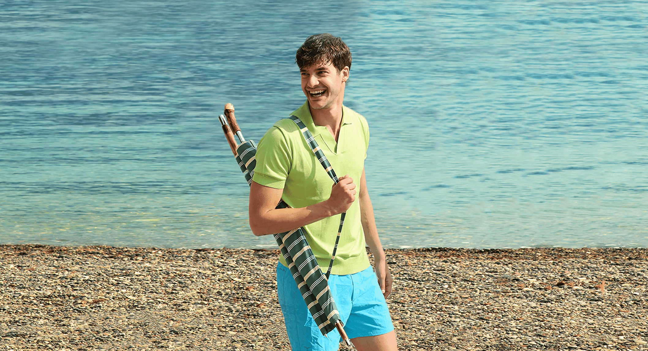 A man walking on a pebble beach wearing a lime green polo shirt and bright blue shorts.