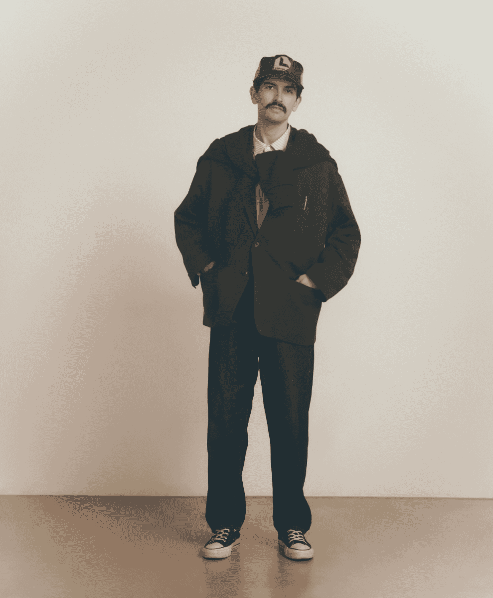 Model in a dark oversized coat and cap standing against a plain backdrop.