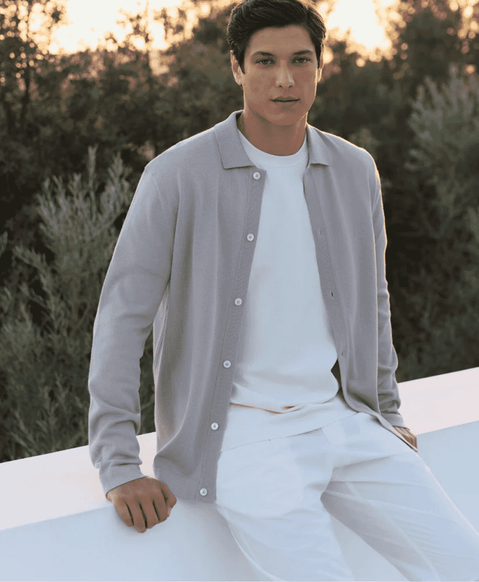 A man in a light gray knitted cardigan and white t-shirt sitting outdoors.