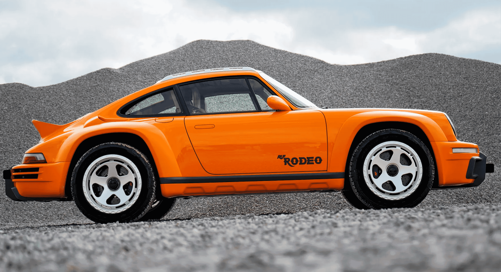 A vibrant orange RUF Rodeo parked on gravel against a backdrop of gray rock piles under a cloudy sky.