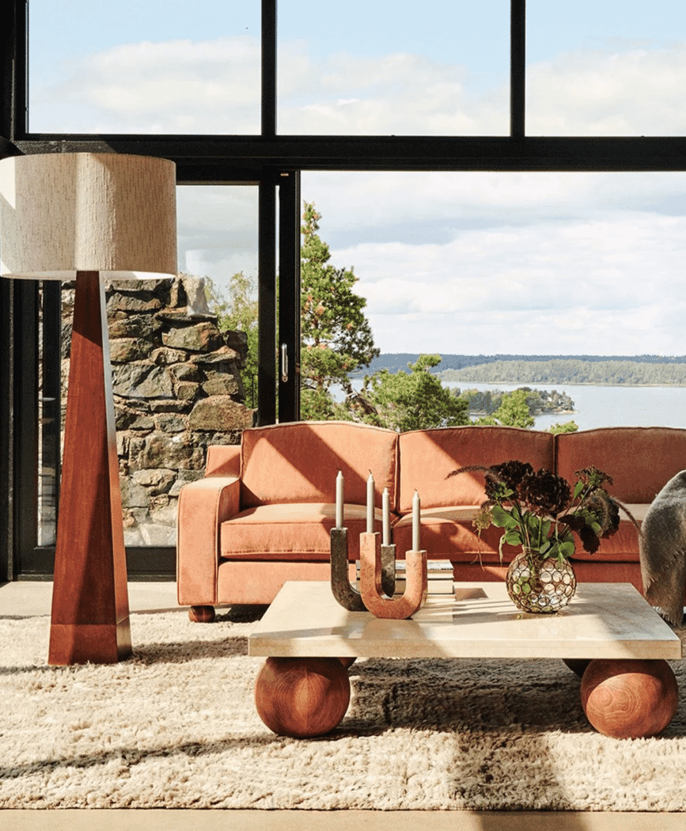 A modern living room with a pyramid-shaped floor lamp beside a stone wall overlooking a scenic lake view.