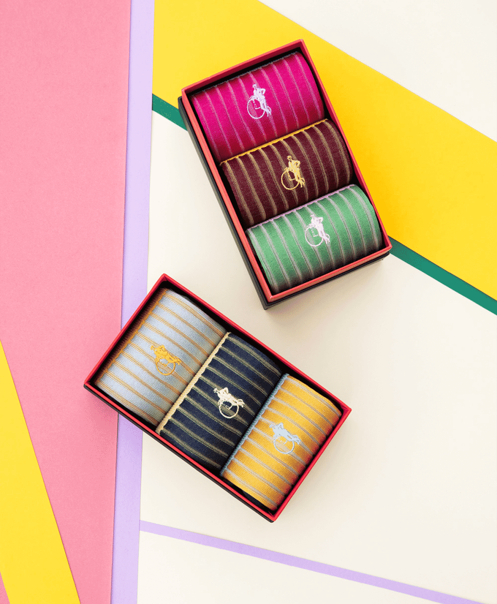 Two gift boxes of striped dress socks on a vibrant pink, yellow, and purple geometric background.