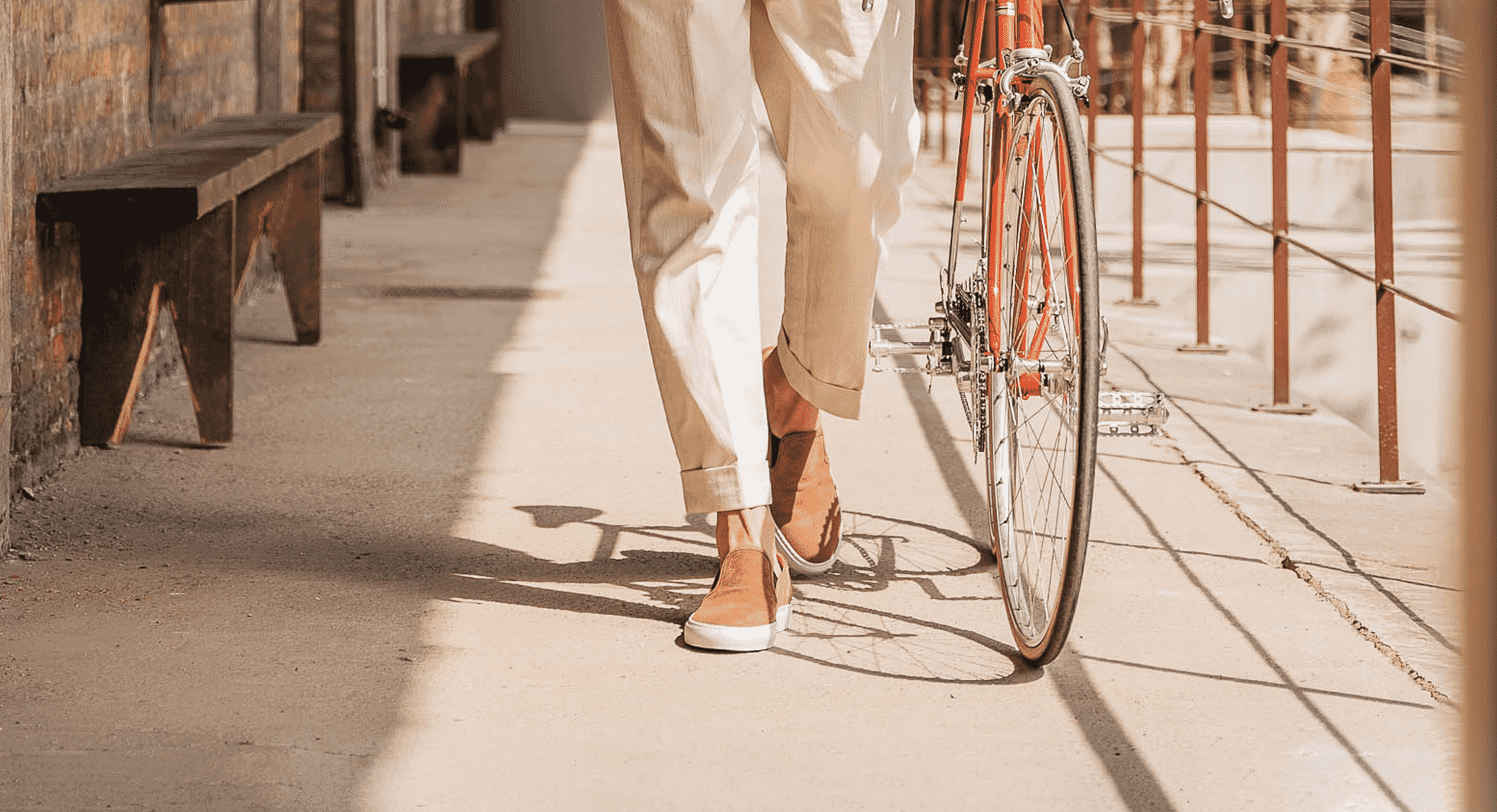 Person in beige trousers and tan slip-on sneakers walking an orange bicycle.
