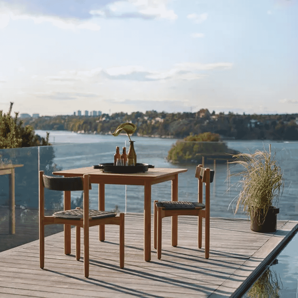 A wooden dining set with woven chairs on a lakeside deck under a clear blue sky.