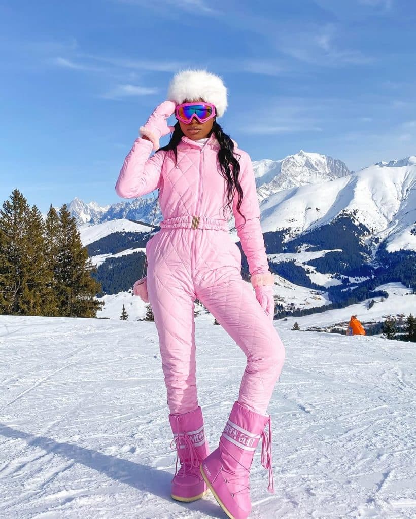 Woman in a vibrant pink quilted ski suit with a fluffy white hat and reflective goggles.
