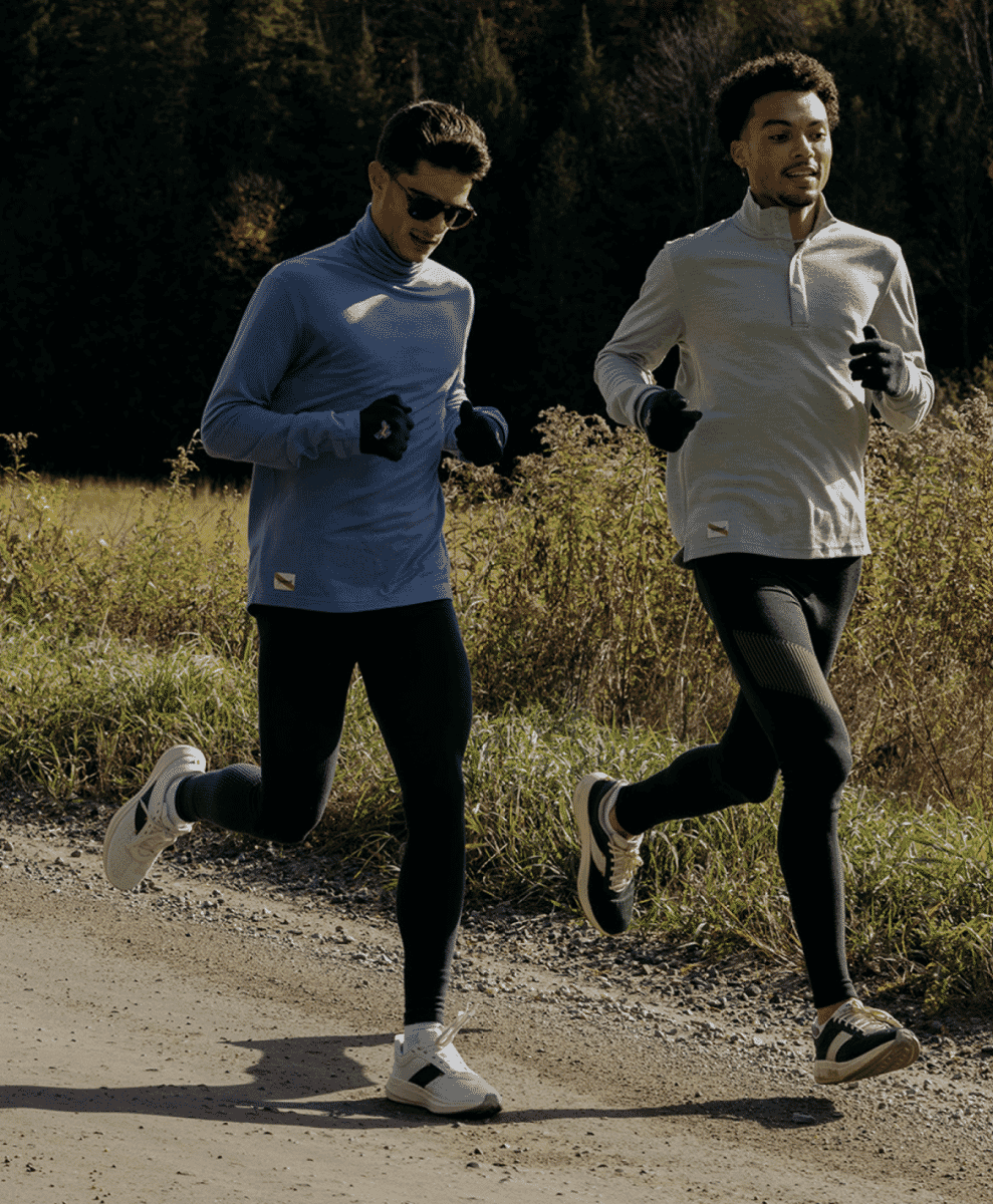 Two men running on a sunlit dirt path surrounded by autumn foliage in technical athletic wear.