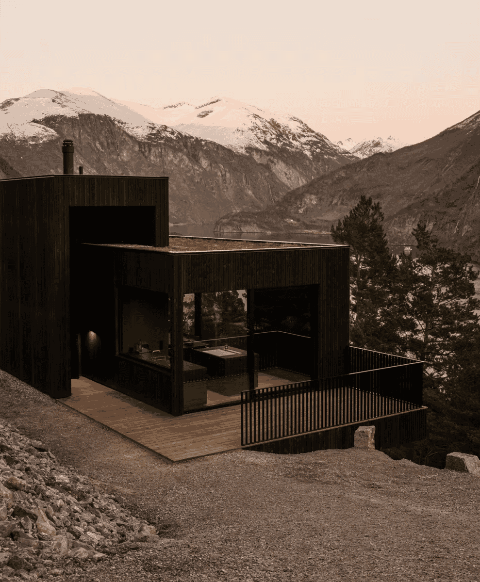 Modern black wooden house perched on a mountain cliff overlooking snow-capped peaks and a fjord.