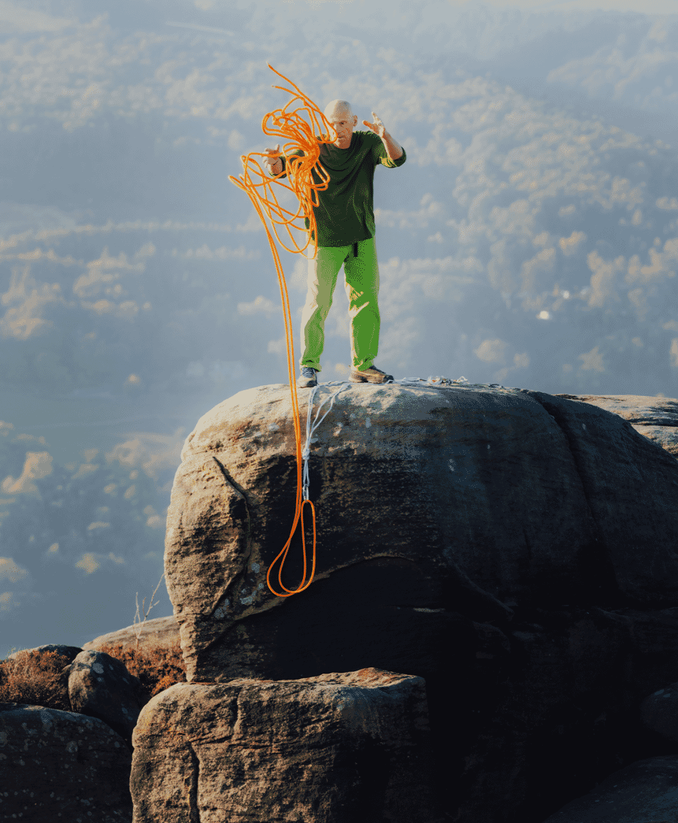 A man in vibrant green pants and top standing on a rocky cliff with climbing rope.