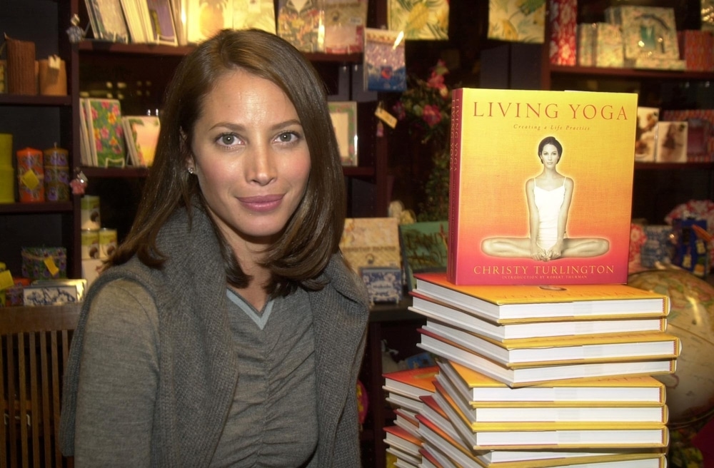 Christy Turlington wearing a gray cardigan standing next to her yoga book 'Living Yoga'.