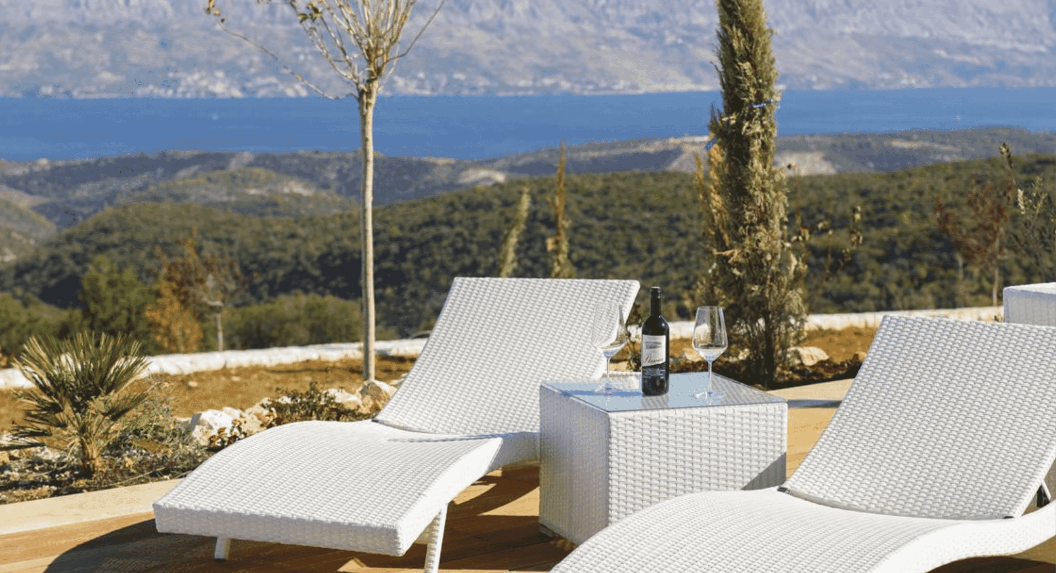Wicker lounge chairs and wine glasses on a terrace overlooking a coastal mountain range.