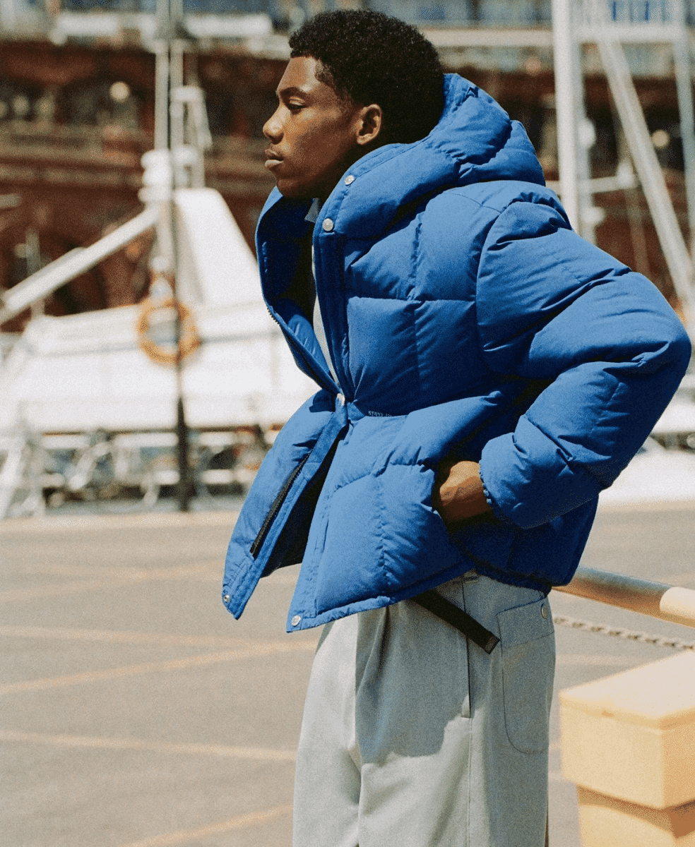 A man in a vibrant blue puffer jacket and gray trousers standing at a marina.