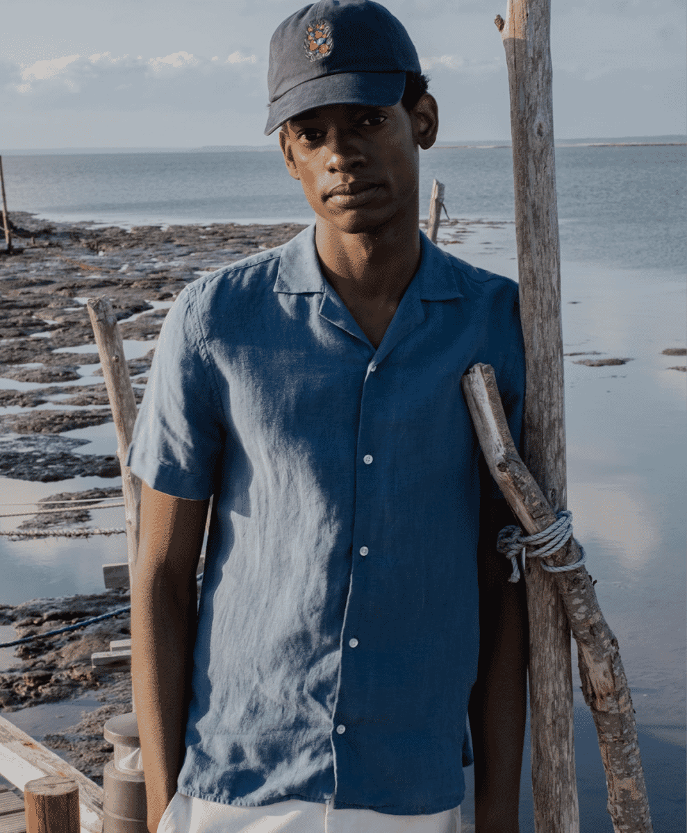 A man wearing a blue linen short-sleeve button-up shirt and a matching cap by a wooden pier.