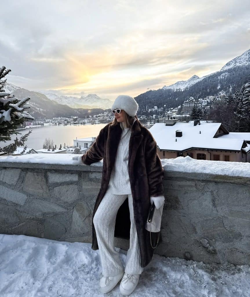 Woman in a brown fur coat against a sunset mountain lake backdrop.