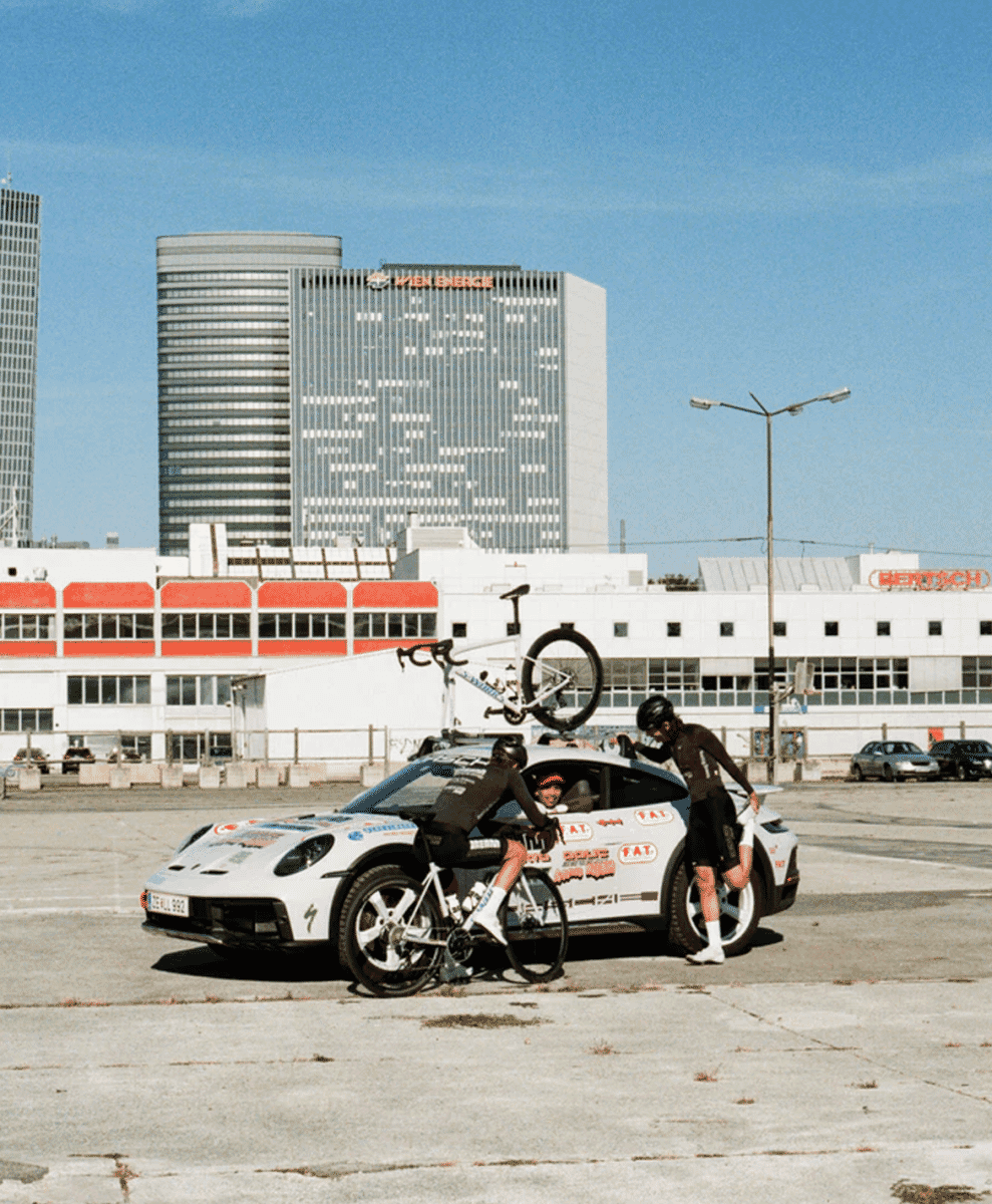 Two cyclists in black cycling kits pose with a white Porsche 911 adorned with sponsor stickers in an urban lot.