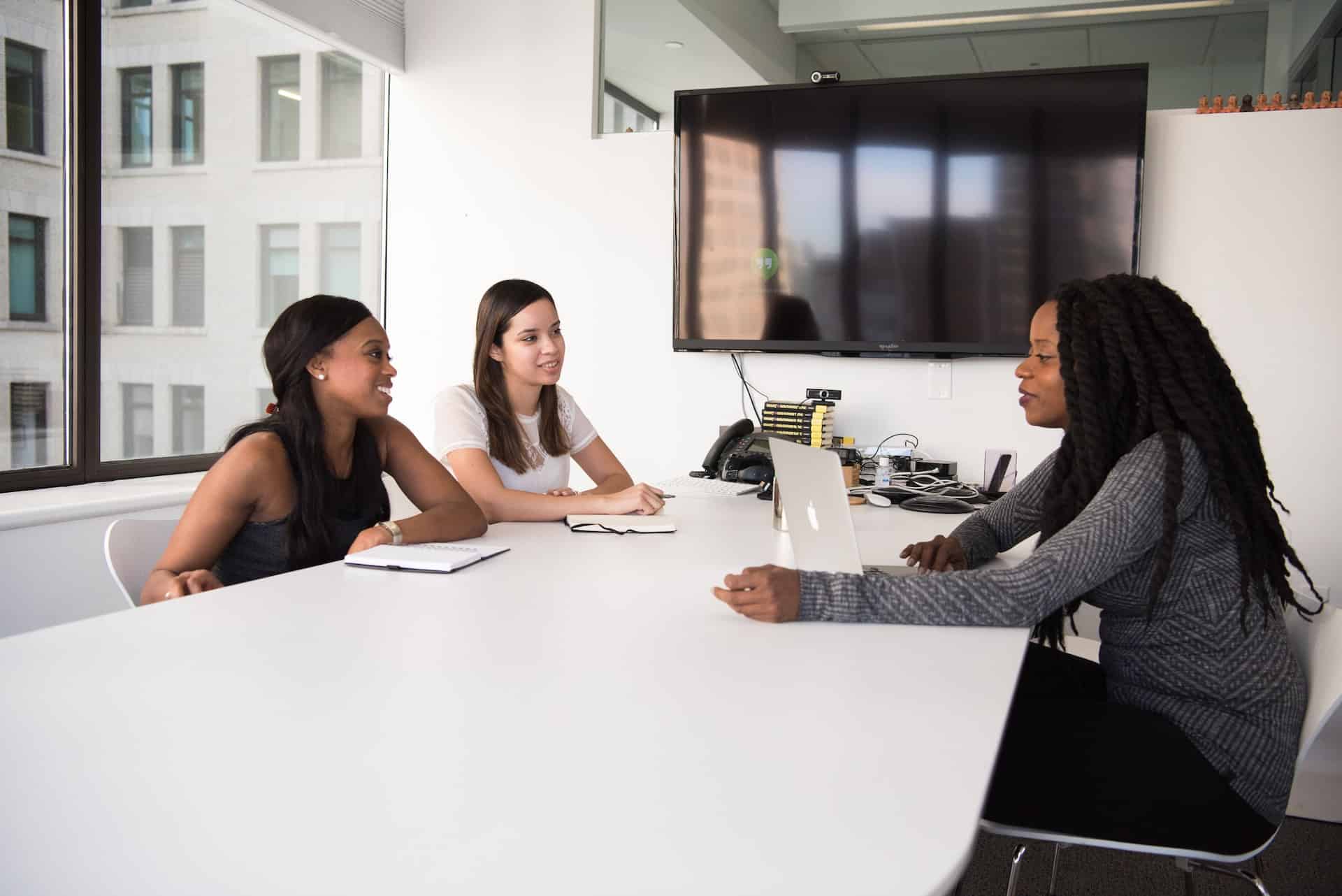 Three professional women in varied business attire working with laptops and notebooks at a modern conference table.