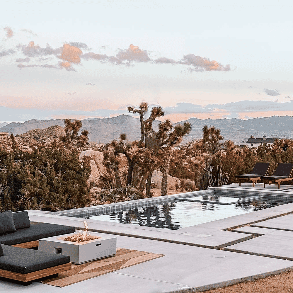 An outdoor patio with modern furniture, a fire pit, and a pool overlooking Joshua trees at sunset.