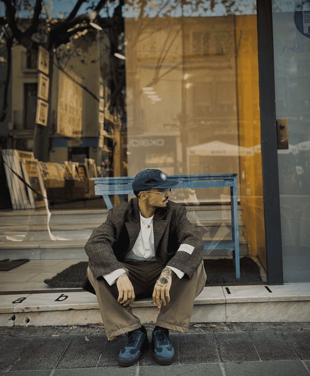 A man in a plaid blazer and beige trousers sitting on a city sidewalk wearing blue sneakers.