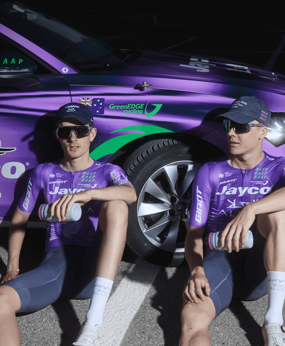 Team Jayco-AlUla riders in matching purple kit and caps sitting in front of a branded team car.