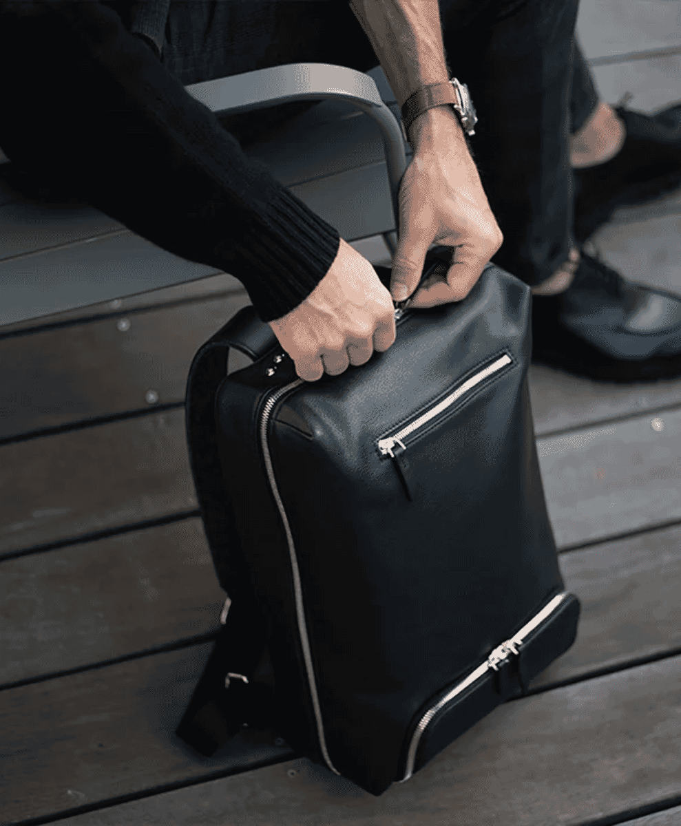 Man in a black ribbed sweater zipping a sleek black leather backpack with silver zippers.