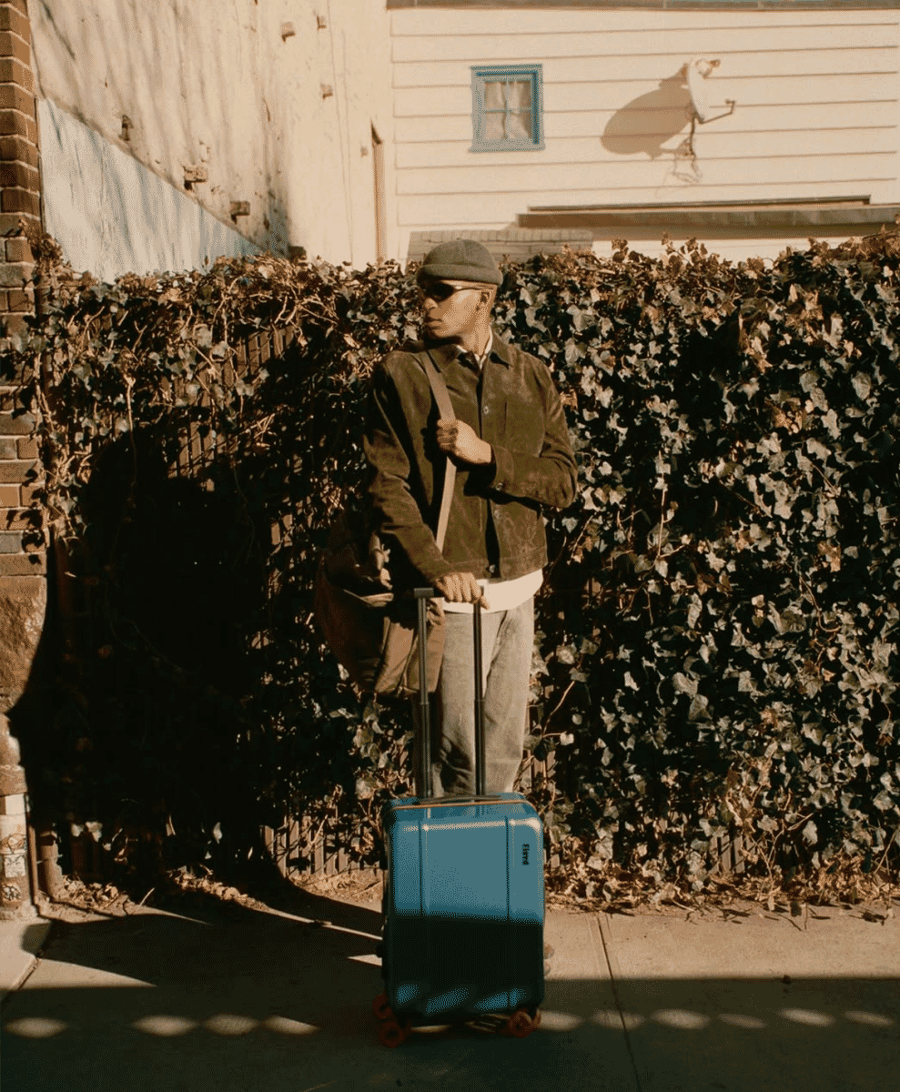 A man in a brown suede jacket pulling a blue carry-on suitcase on an urban sidewalk