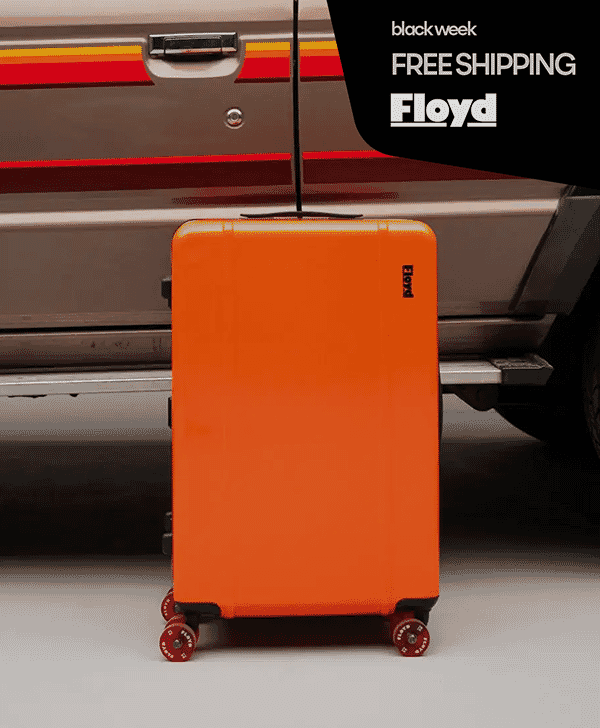 An orange Floyd hard-shell suitcase in front of a vintage vehicle.