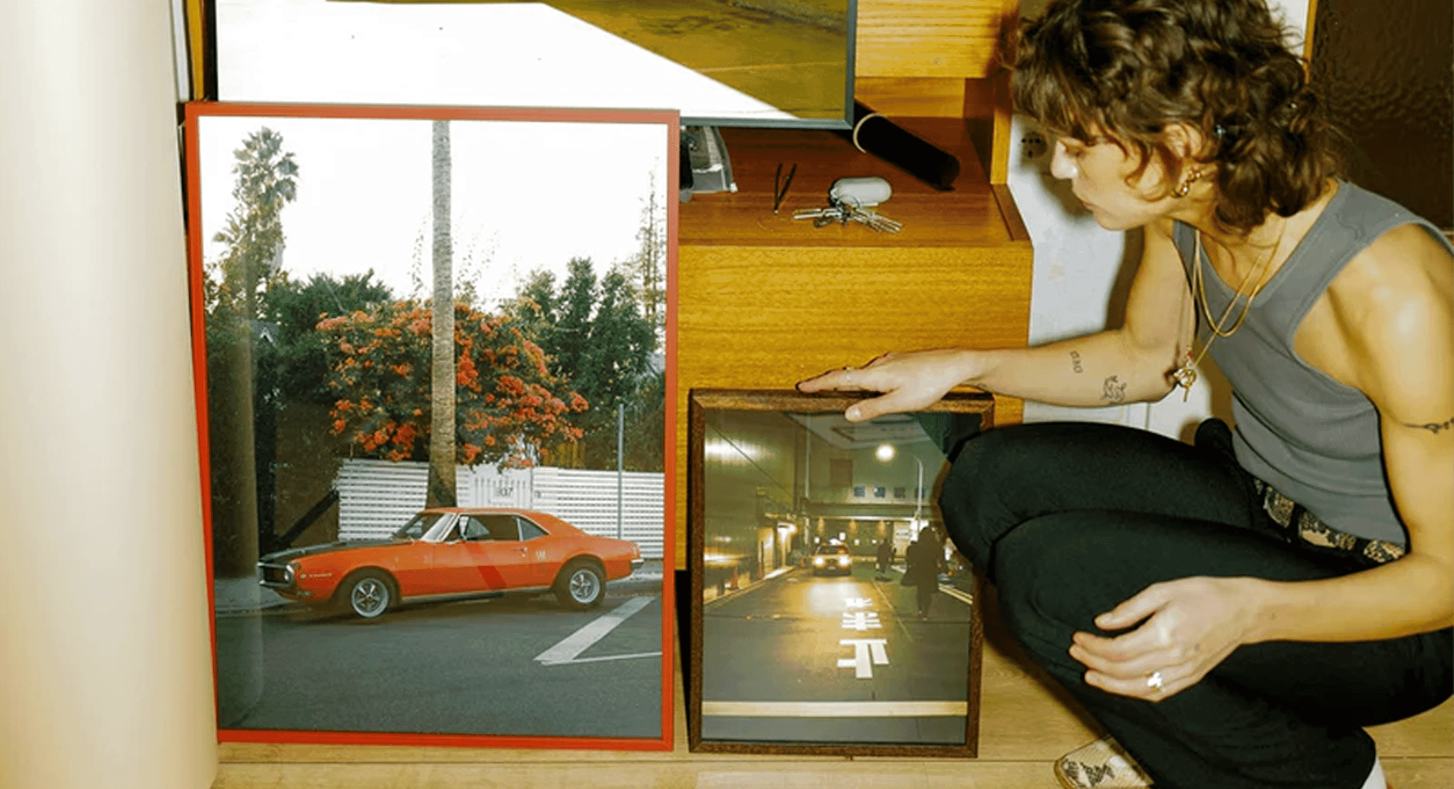 A person in a gray tank top and black pants crouching beside framed photos and vintage cameras in a wooden interior.