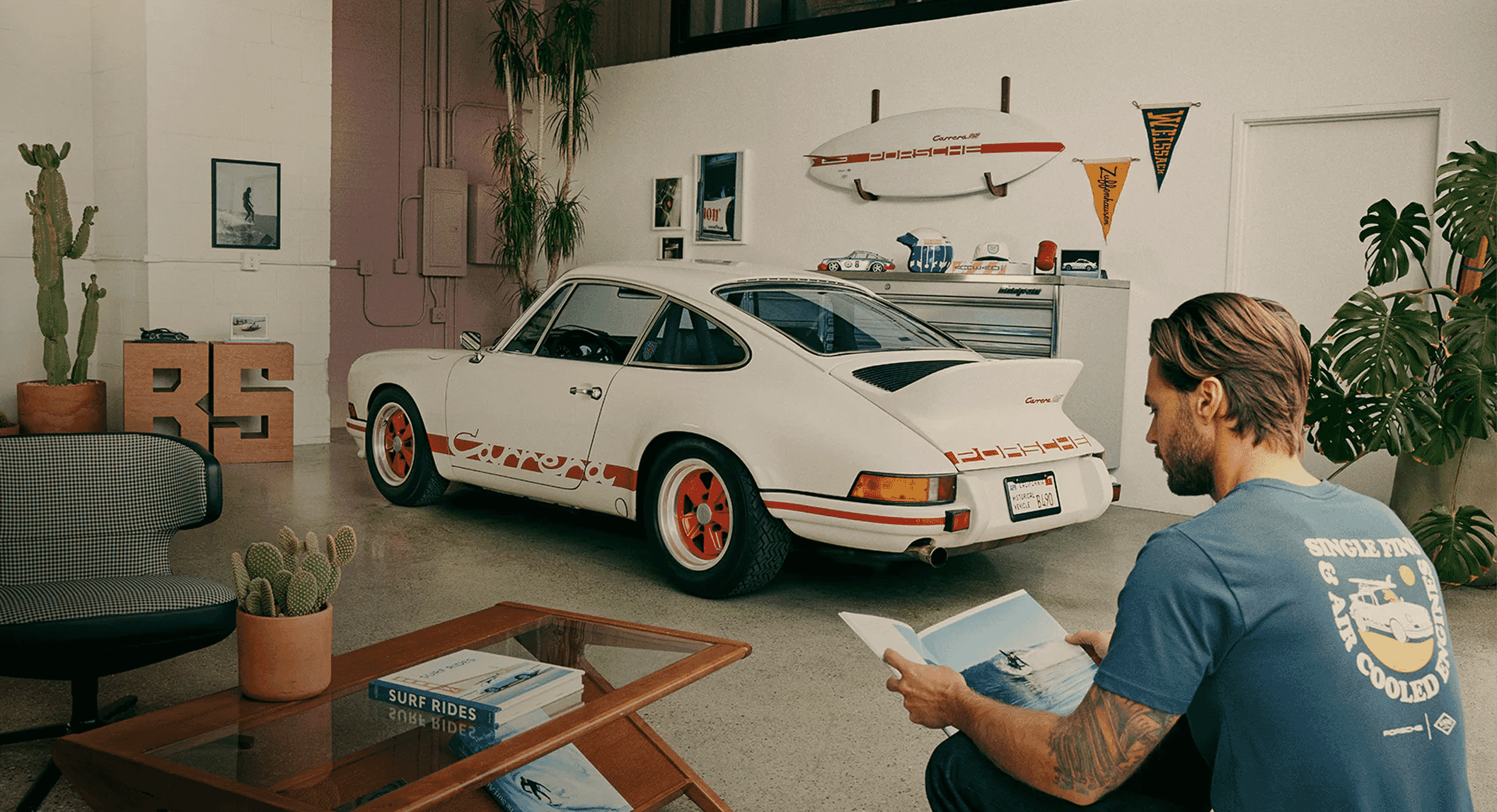 A man in a blue graphic t-shirt sitting in a garage with a white Porsche 911 and surfboards.