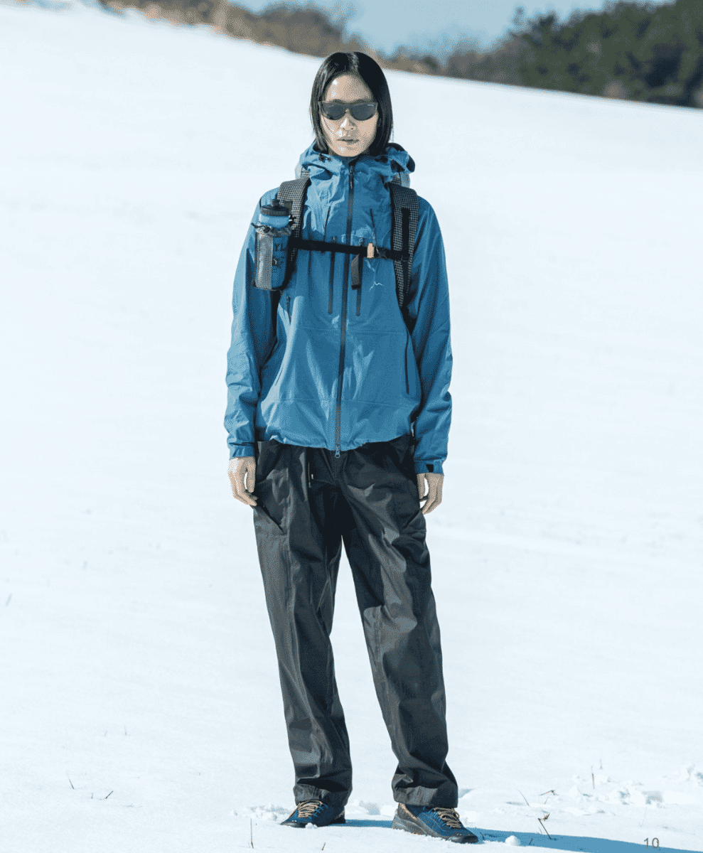 A person in a blue technical jacket standing on a snowy slope looking toward the horizon
