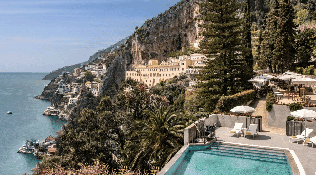 A luxury resort poolside with lounge chairs overlooking a cliffside Mediterranean coastal town.