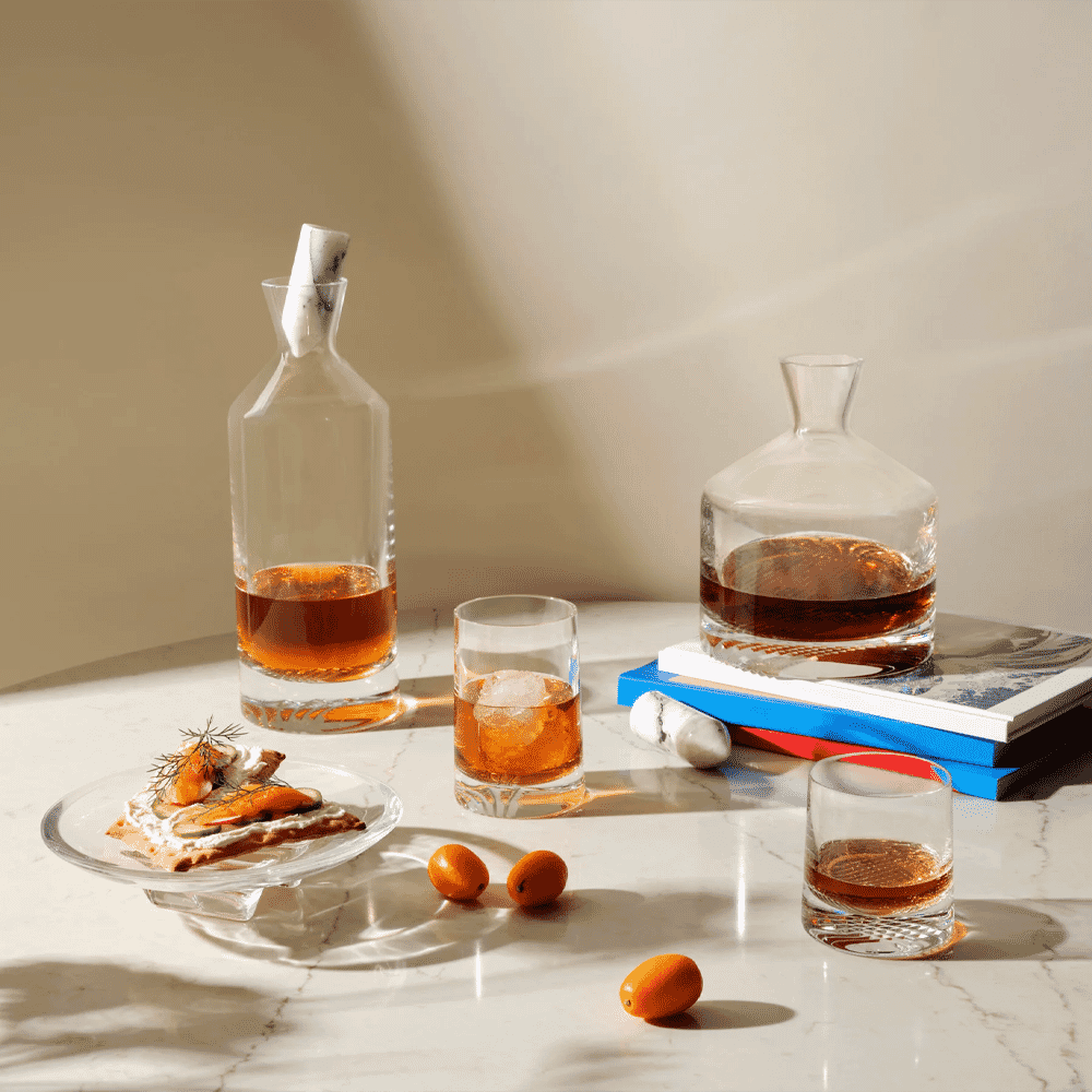A luxury still life with whiskey decanters, glasses, and kumquats on a marble table.