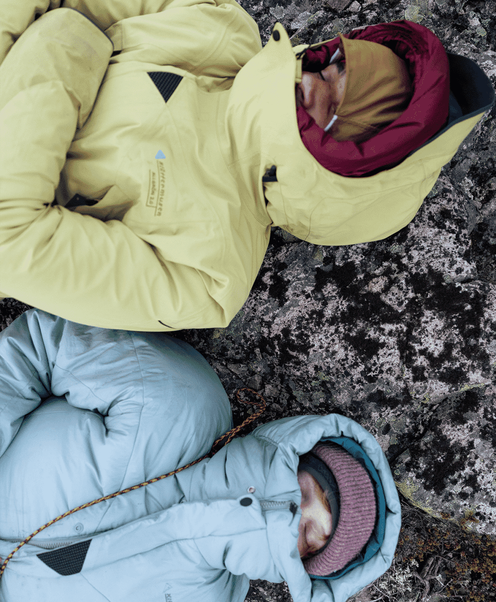 Two people resting on rocky terrain wearing insulated hooded technical gear.