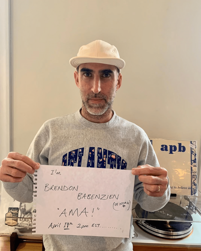 Man in a floral sweatshirt holding a handwritten sign in an indoor setting.
