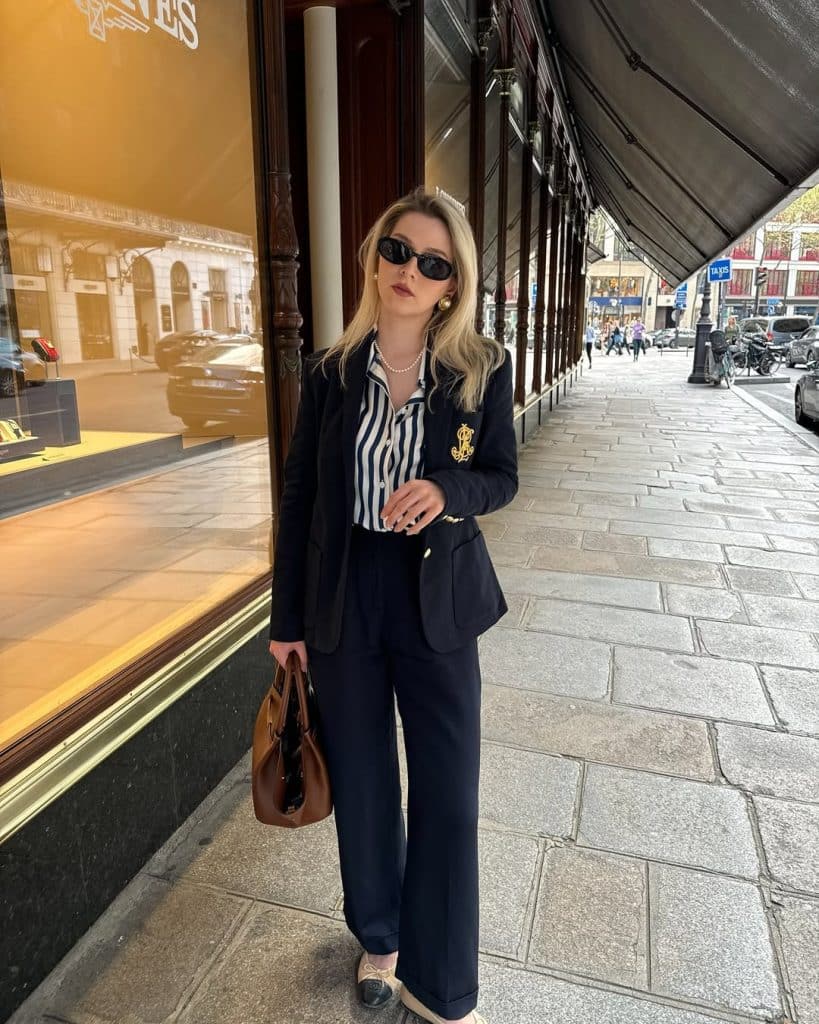 A woman in a navy blazer with gold crest embroidery and a striped shirt walking on a city street.