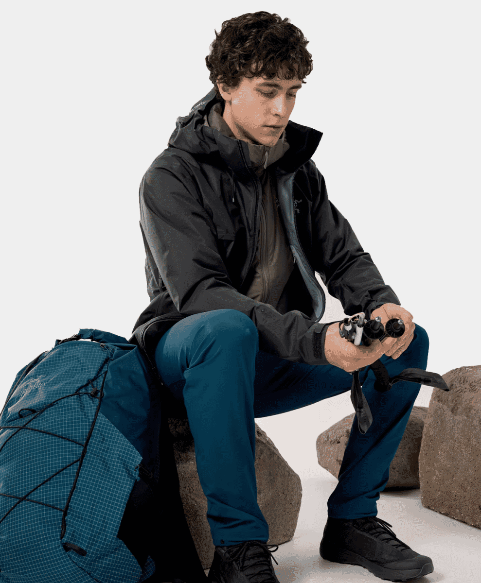 Man in outdoor technical gear sitting on rocks with trekking poles and backpack.