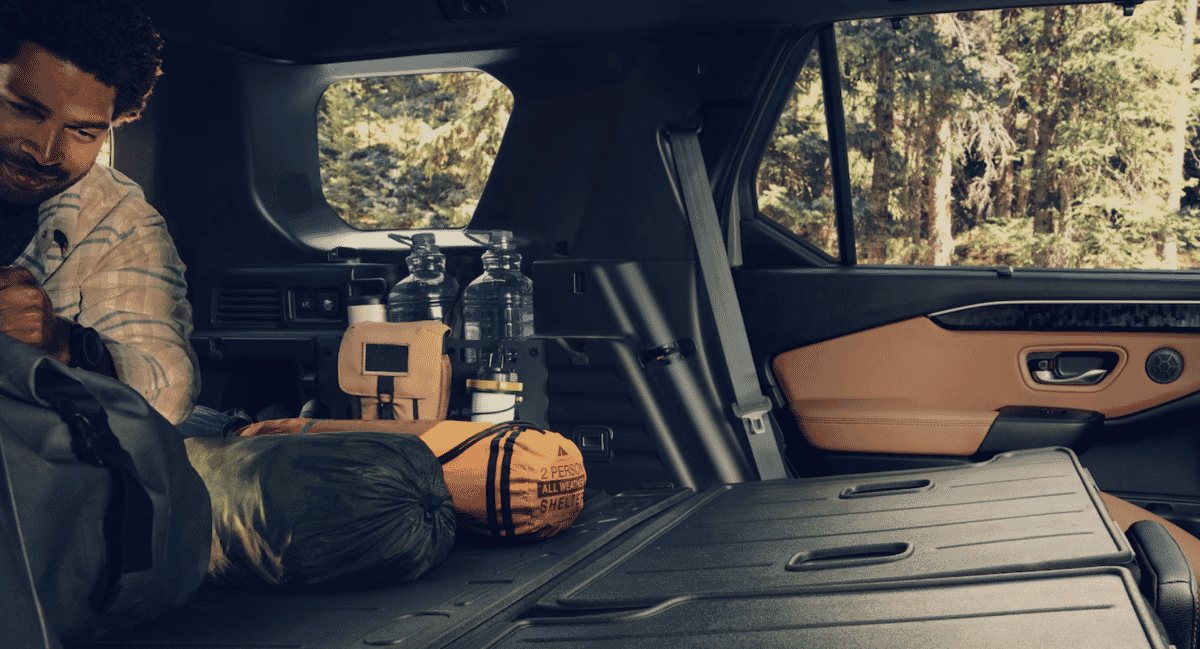 A man in a plaid shirt sits in the back of an SUV packed with camping gear.