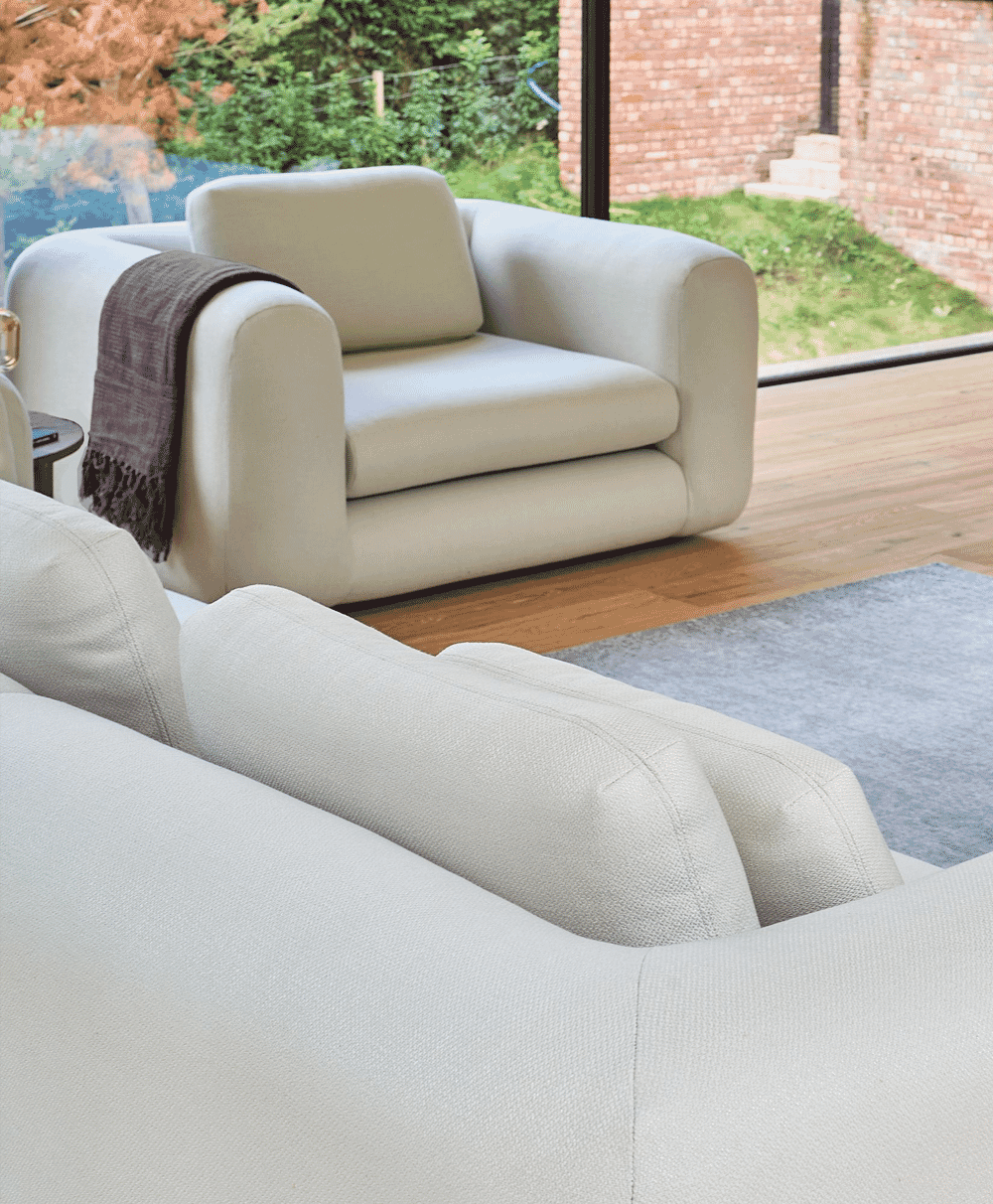 Beige upholstered armchair and sofa in a bright living room with wooden floors and outdoor greenery.